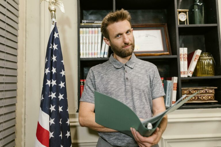 Business professional standing confidently in office holding documents near American flag.