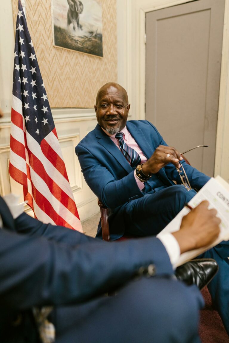 Two professionals in discussion by an American flag in an office setting.